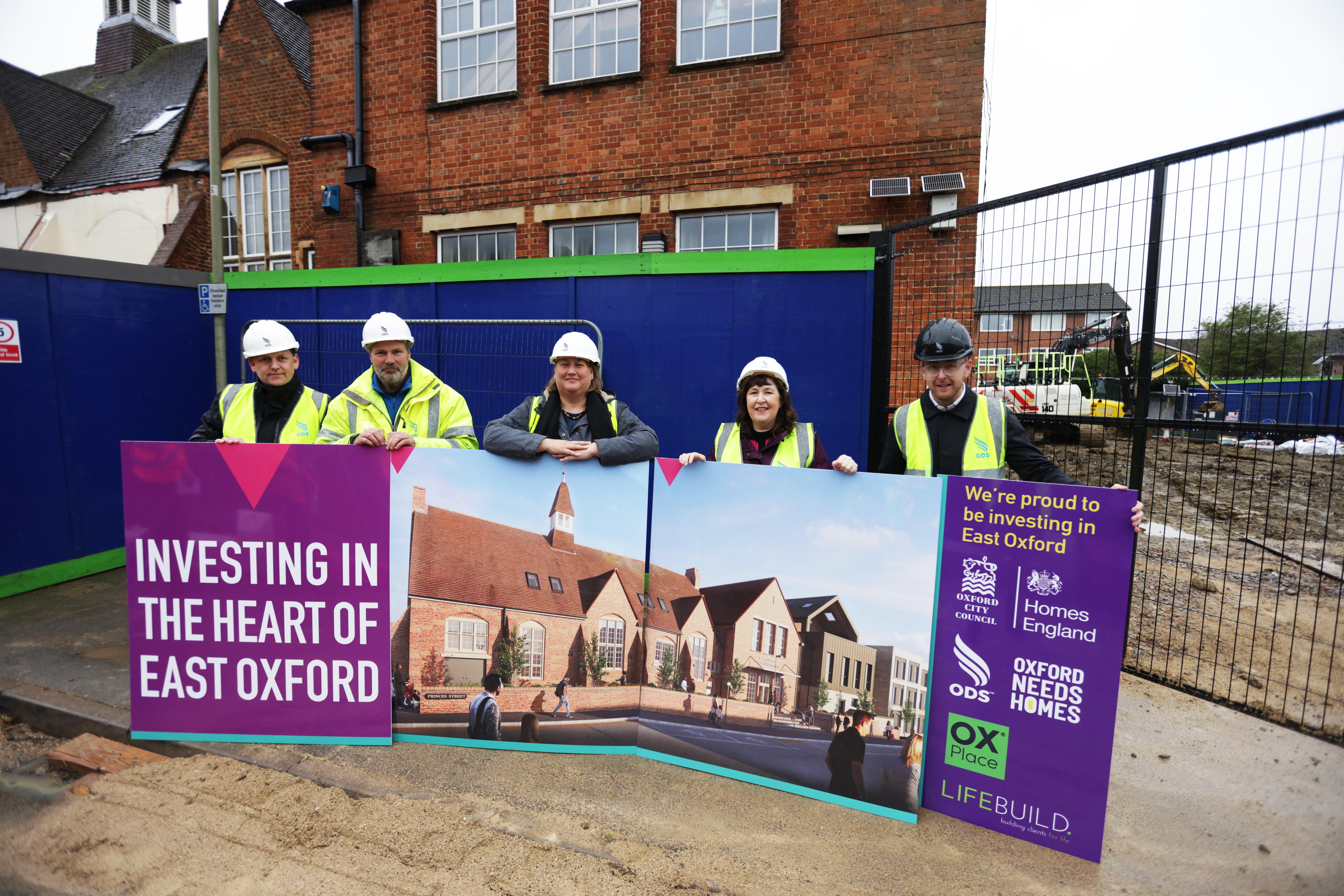 A group of five individuals in his-visibility yellow jackets and white hard hats standing in front of the brick East Oxford Community Centre building. They are holding some large signs which show what the building will look like when work is completed.