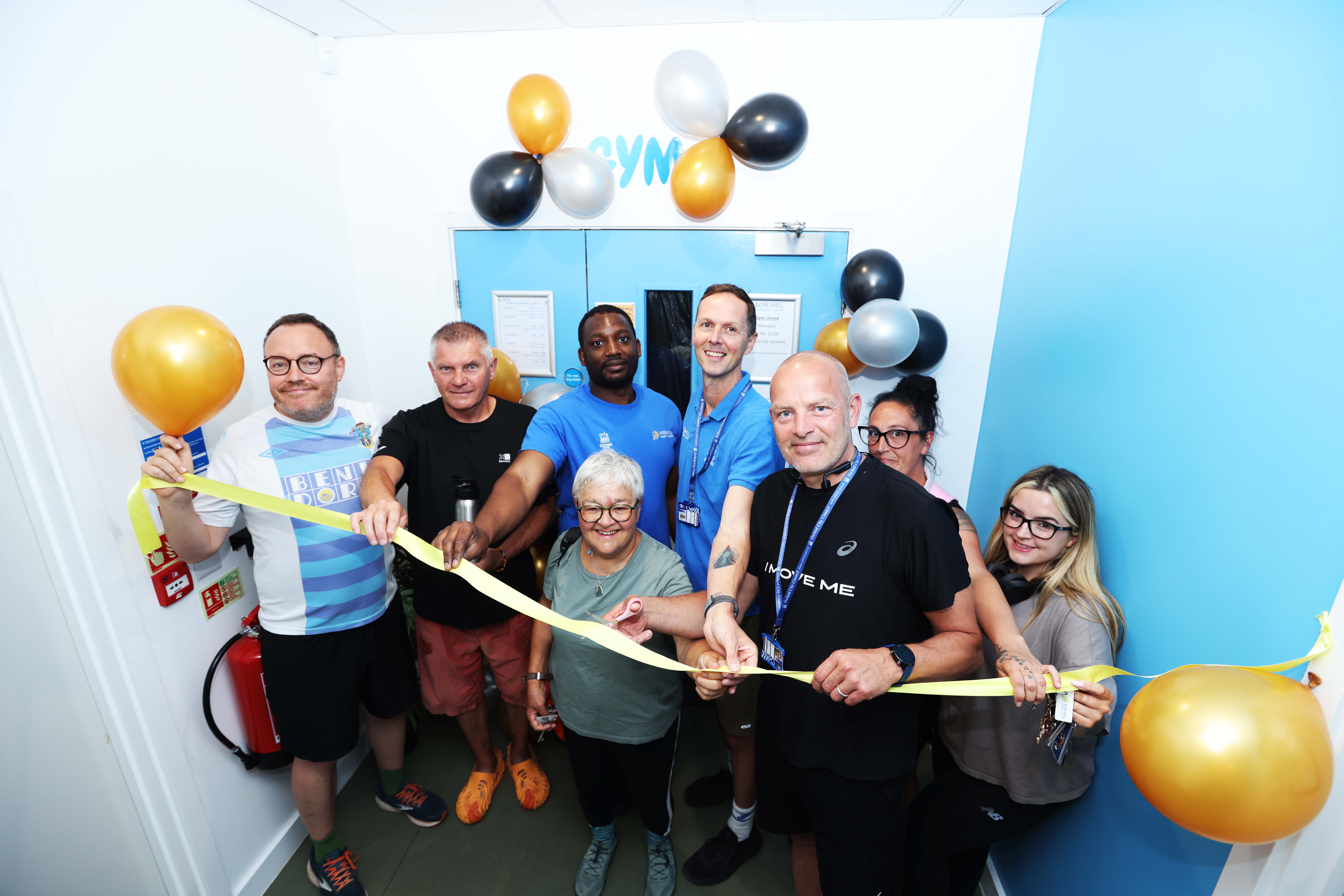 Councillors Mark Lygo and Ed Turner are joined by centre staff and users holding balloons and cutting a ribbon to mark the reopening of the gym